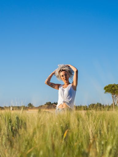 An adult woman with white curly hair and tattoos posing in a field on a sunny day in the countryside