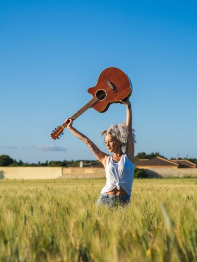 A cute Spanish woman with white curly hair carrying a guitar above her head in a field during sunset