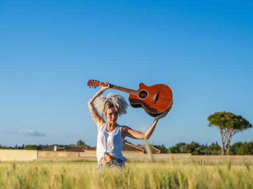 A cute Spanish woman with white curly hair carrying a guitar above her head in a field during sunset