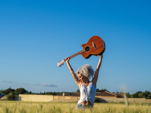 A cute Spanish woman with white curly hair carrying a guitar above her head in a field during sunset