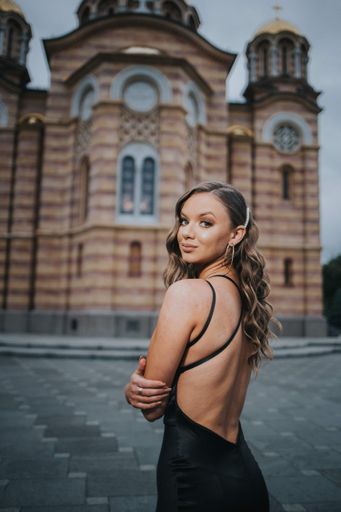 A vertical shot of a Caucasian sexy woman with a black evening dress on the Cathedral of Christ the Saviour Banja background