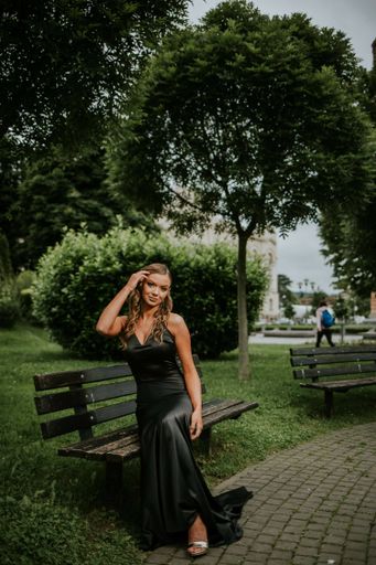 A full body shot of a beautiful elegant woman in a fancy black dress sitting on a bench in the park