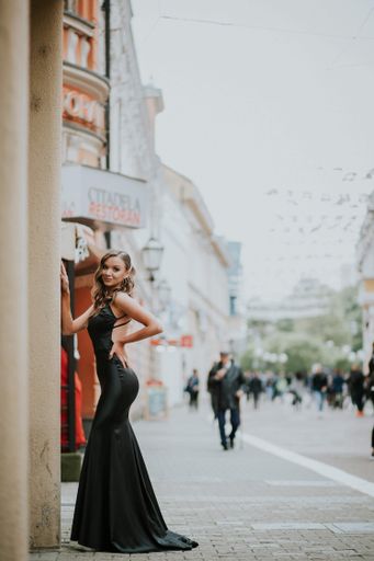 A vertical shot of a Caucasian sexy woman with a black evening dress leaning on the street columns