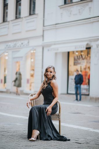 A full body shot of a beautiful elegant woman in a fancy black dress sitting on chair outside