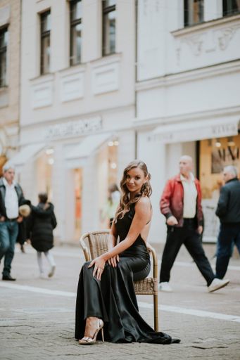 A shallow focus of a young elegant woman wearing a long black dress and sitting on a chair on the street
