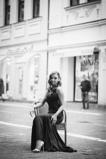 A vertical shot of a Caucasian woman with a black evening dress sitting on a chair in the street