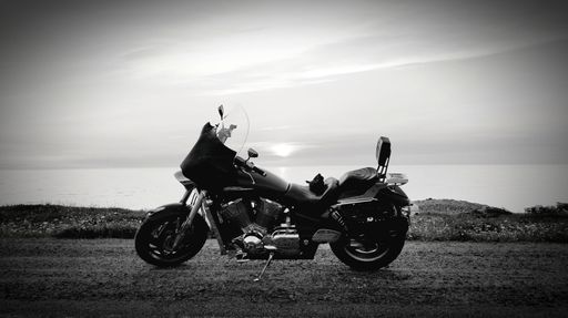 A black and white photograph of a motorcycle parked on the shore next to a body of water.