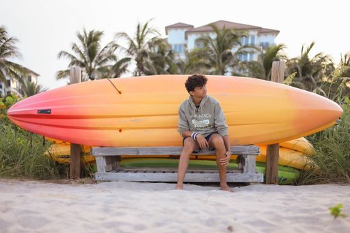 A young adult man in a relaxed pose seated on a park bench, with a collection of colorful surfboards at his feet