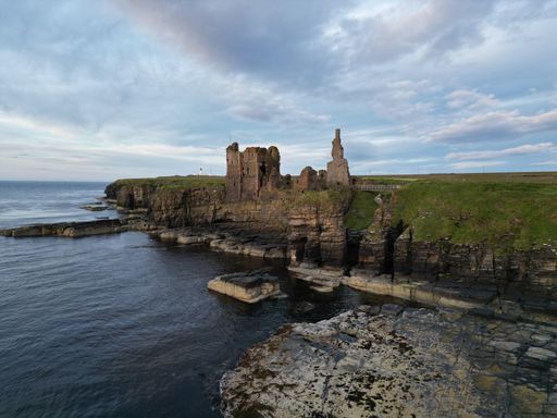 An aerial view of the historic Sinclair Girnigoe Castle ruins on a rocky outcrop at sunset