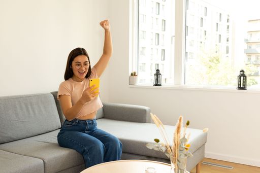 a woman sitting on the sofa at home, happily raising her arm while looking at the cell phone