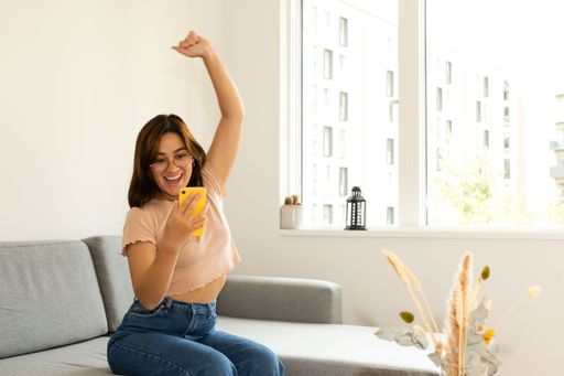 a woman sitting on the sofa at home, happily raising her arm while looking at the cell phone