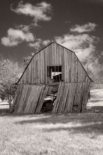 A black and white vertical shot of an old wooden barn leaning to one side under cloudy skies