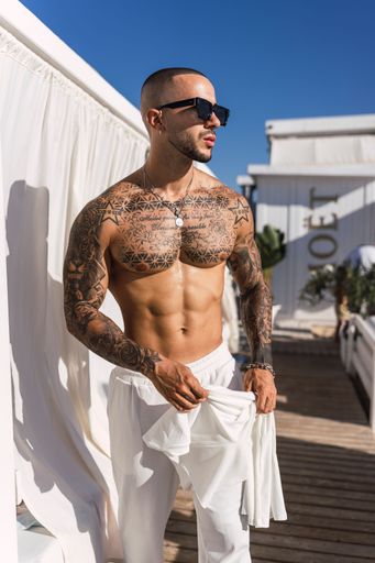 A young, shirtless man with tattoos on a deck posing for the camera