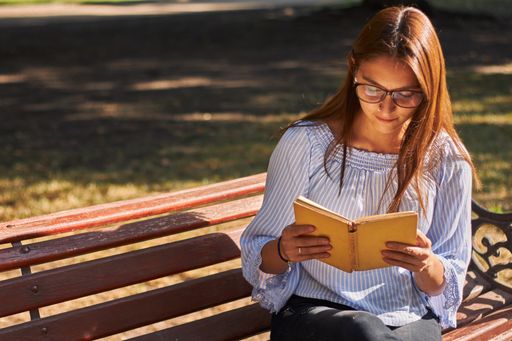 A beautiful shot of a girl in a blue shirt and with glasses reading a book on the bench