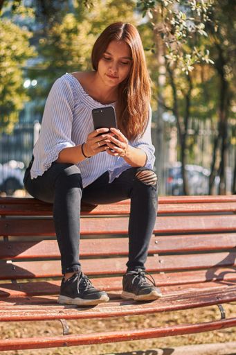 A vertical shot of a girl sitting on the bench and looking at her phone