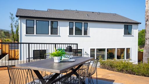 A scenic outdoor patio setting featuring a wooden table and four chairs, with the backdrop of a house and surrounding greenery