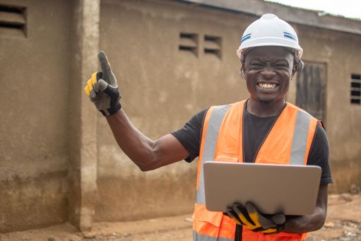 A male construction worker wearing a safety vest and a bright yellow construction vest is looking intently at his laptop computer