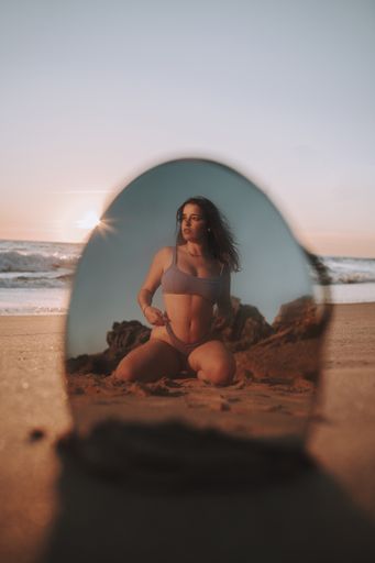 A vertical shot of a sexy Spanish Caucasian woman posing in a bikini on a sandy beach, photographed through a mirror