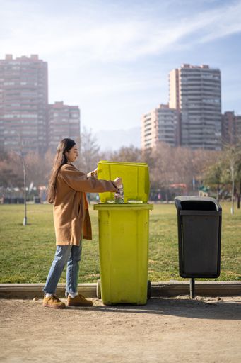 A young woman throwing a plastic water bottle into a green, public park trash can