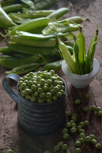A vertical shot of a heap of emptied pea pods and a metal basket full of peas on a wooden table