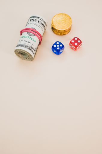 A vertical shot of rolled dollars with a red rubber band, gold coins, and dice on a cream-colored surface