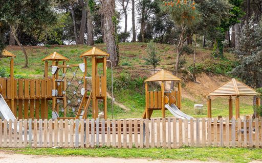 A wooden playground on the sand in the fenced-in park at the foot of a hill on a sunny day