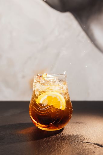 An artistic image of a clear glass filled with a bubbly, yellow-hued alcoholic beverage, garnished with small slices of lemon
