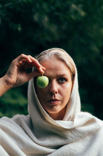 A vertical shot of a female in a shawl holding an apple recreating the "Son of a Man" painting