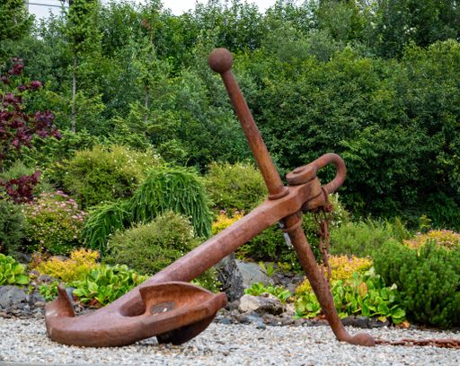 An aged anchor rests atop a bed of gravel in a natural outdoor setting, surrounded by trees