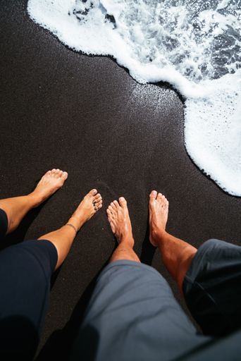 A high-angle view of a couple's feet on the black sand beach with soft foamy waves