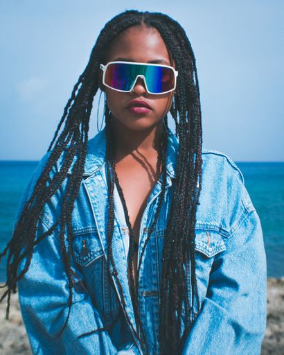 A vertical shot of a black young lady wearing big sunglasses with ocean in the background