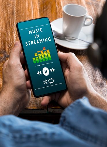 The hands of a man holding on a wooden table a smartphone playing streaming music. "Music in streaming" on the smartphone screen