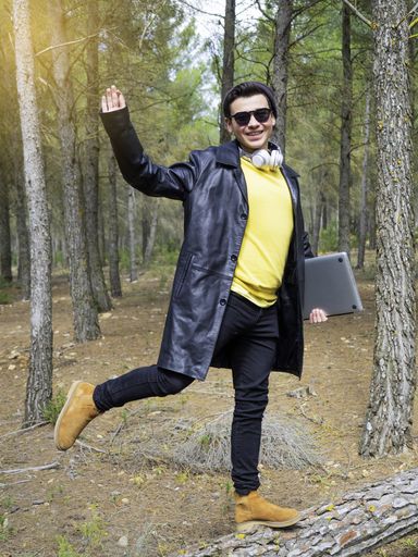 A happy teenager wearing a cap and sunglasses holding a laptop computer and standing in a park