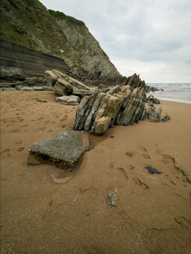 A vertical shot of a sandy beach with a cliff on the shore and big rocks in the foreground