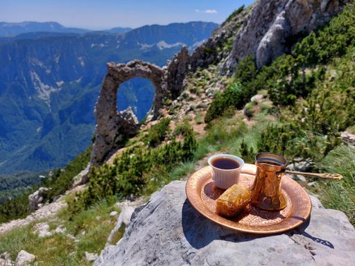 A white coffee cup with a matching coffee pot placed atop a rocky mountain peak with a bright blue sky in the background