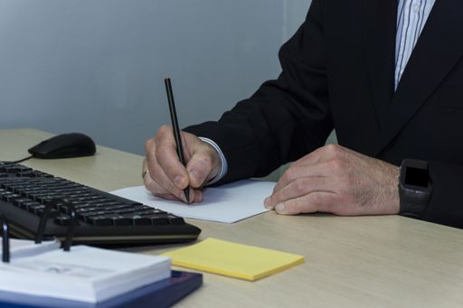 A closeup shot of male writing on paper with a computer keyboard and office supplies on the desk