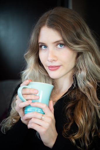 Attractive blonde woman wearing a black top and blue eyes looking directly at the camera while holding a blue ceramic cup in her hands