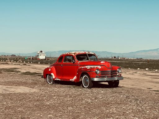 A vintage red car parked in a field, surrounded by tall mountains