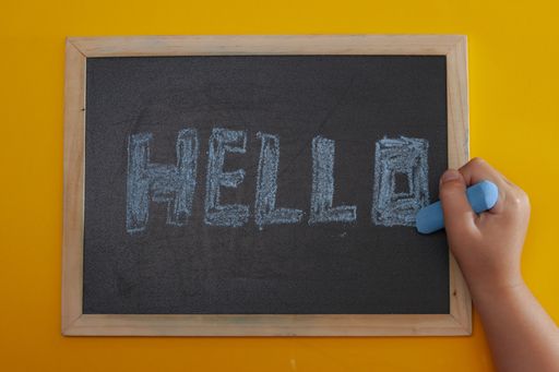 A closeup shot of a board with the word hello written in chalk in blue with a girl's hand
