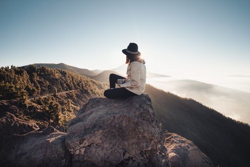 A panoramic shot of a female with a hat sitting backwards on the edge of a cliff  with a mesmerizing view of foggy hills