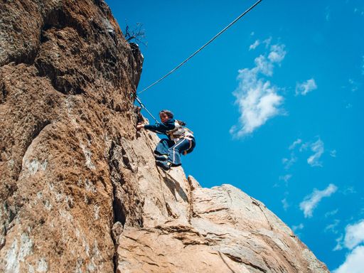 A young adult male is scaling a steep and large rock face, as he reaches for the top with a determined expression