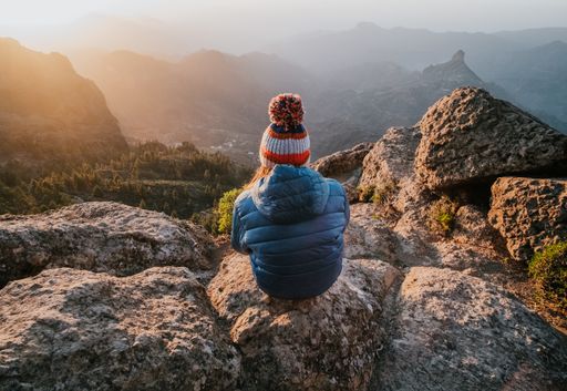 A beautiful panoramic shot of a mesmerizing view of rocky mountains from the top and a female with a beanie sitting backwards on a stone