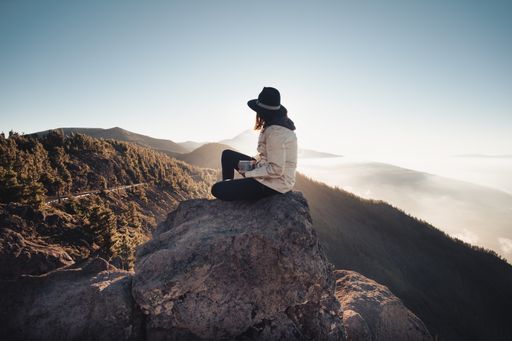 A panoramic scenery of rocky mountains covered in fog and a female with a hat sitting backwards on the edge