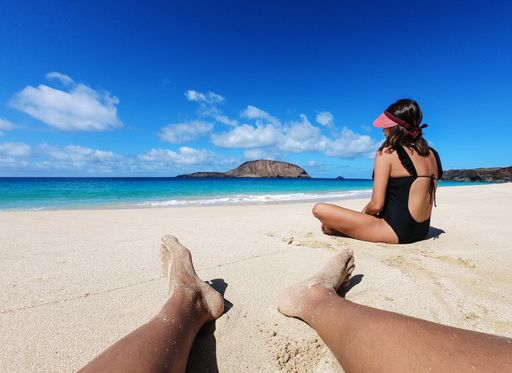 A POV shot of a sunny tropical sandy beach with a blue sky and a female in a swimsuit sitting nearby in Playa De Las Conchas Casas, Spain