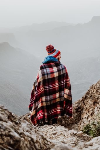 A woman with plaid poncho and pompom beanie hat overlooking a beautiful mountain landscape in the morning