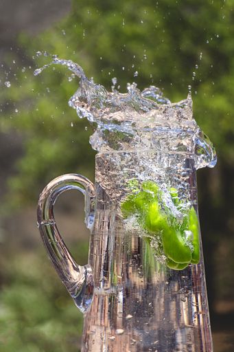 A green pepper being submerged into a glass jug outside while causing big splashes of water
