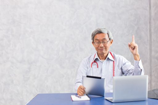 An old, happy male doctor sitting at a desk, using laptop