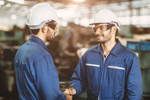 Two male industrial workers wearing protective hard hats and work overalls shake hands in a working environment