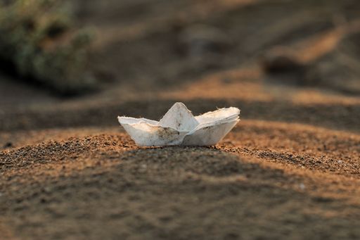 Closeup of a handmade white paper boat resting on a sandy beach, with a blurred oceanic landscape in the background