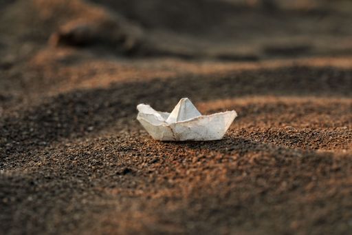 Closeup of a handmade white paper boat resting on a sandy beach, with a blurred oceanic landscape in the background